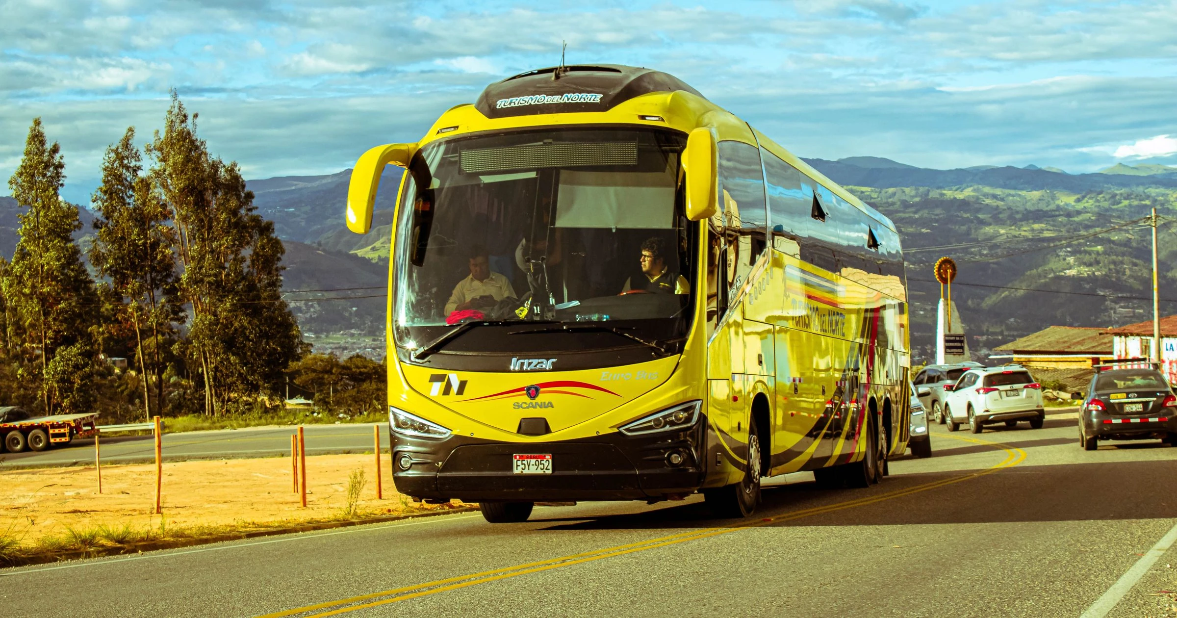 A bright yellow bus travels on a scenic mountain road under a blue sky, surrounded by cars and nature.