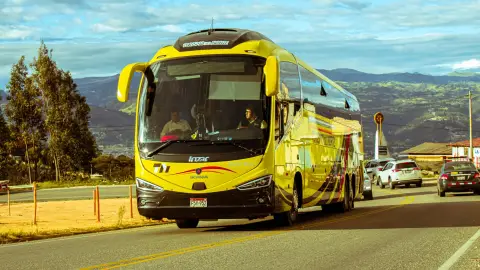 A bright yellow bus travels on a scenic mountain road under a blue sky, surrounded by cars and nature.